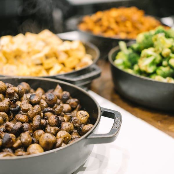 Steaming cast iron pots filled with roasted mushrooms, potatoes, and broccoli on a buffet.