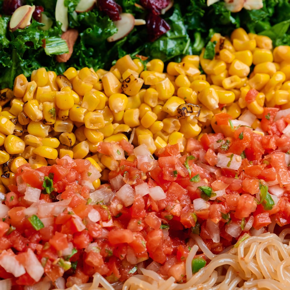 Close-up of a colorful salad with rows of kale, corn, pico de gallo, and noodles.
