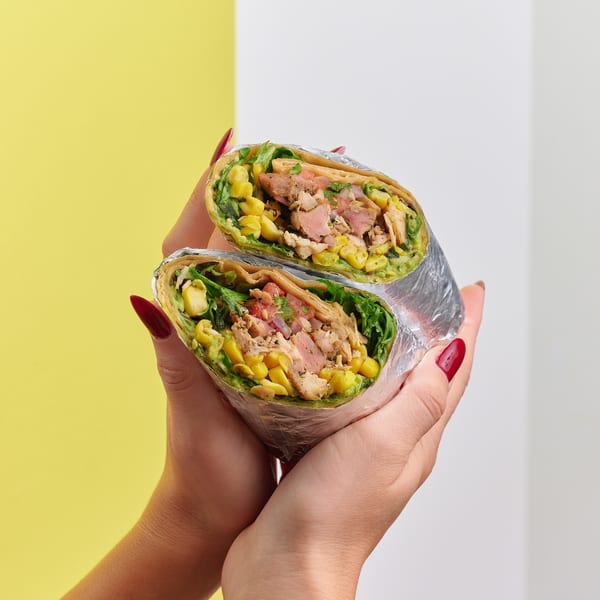 Hands with red nails holding a foil-wrapped burrito cut in half, revealing chicken, corn, and greens.