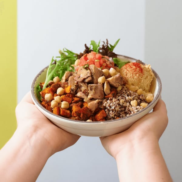 Hands holding a colorful bowl filled with chicken, quinoa, chickpeas, sweet potatoes, hummus, and greens.