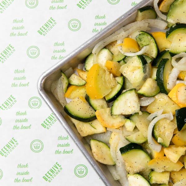 Chopped zucchini, yellow squash, and onions seasoned with pepper in a metal pan on Beehive Kitchen paper.