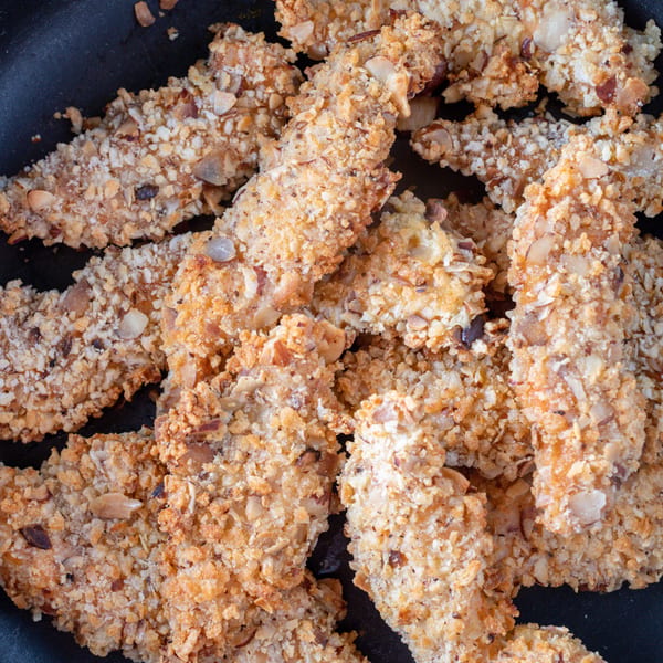 Close-up of golden-brown, nut-crusted chicken tenders in a black pan, ready to be served.