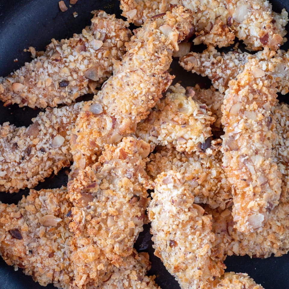 Close-up of golden-brown, nut-crusted chicken tenders in a black pan, ready to be served.