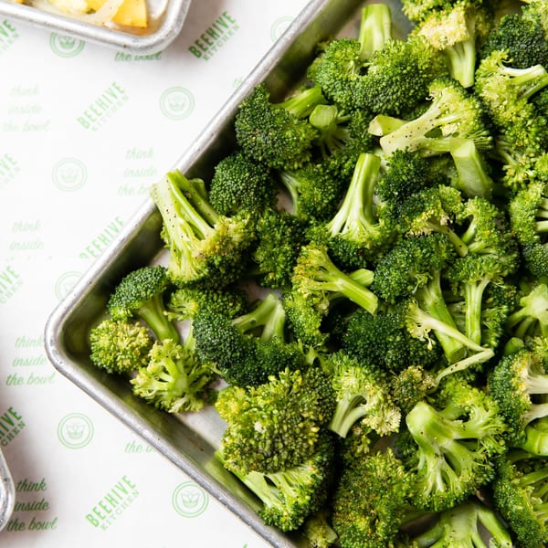 Fresh green broccoli florets seasoned with pepper in a metal tray at Beehive Kitchen.