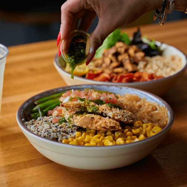 A hand pours green sauce over a colorful bowl of quinoa, corn, green beans, and chicken.