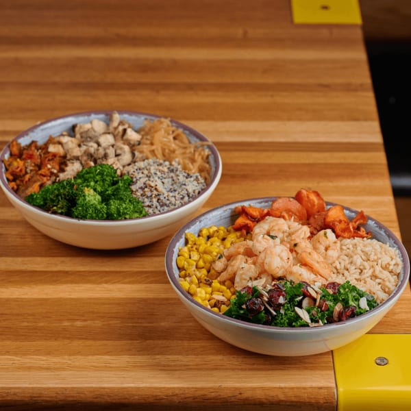 Two colorful bowls of food on a wooden table, one with shrimp and the other with chicken.