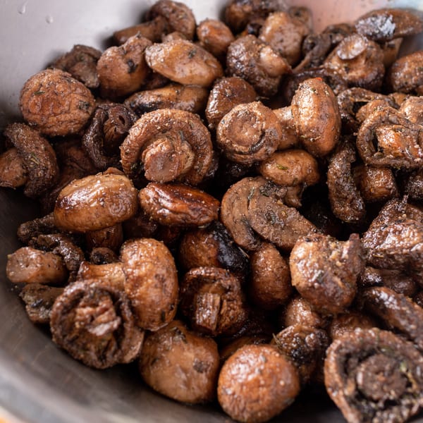 Close-up of a metal bowl filled with cooked, seasoned mushrooms on a wooden surface.