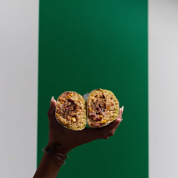 A hand holds a large burrito cut in half, revealing rice, corn, and meat filling.